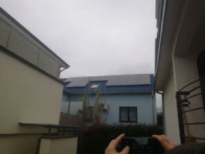 žena držící mobil a fotí modrý dům se střechou na které je fotovoltaika